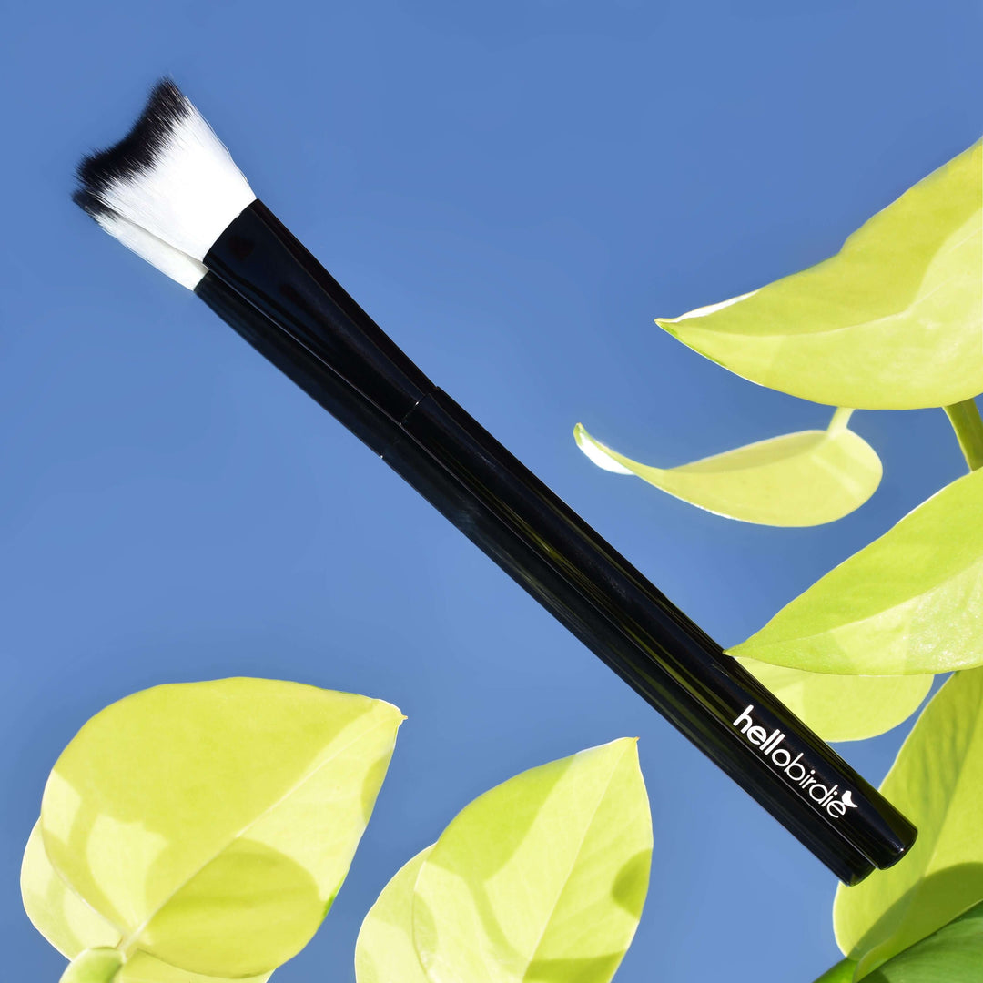 Lash cleansing brush sitting on a mirror with vibrant green leaves and clear blue sky in the reflection. The brush has a shiny black handle with the Hello Birdie logo in white toward the base.