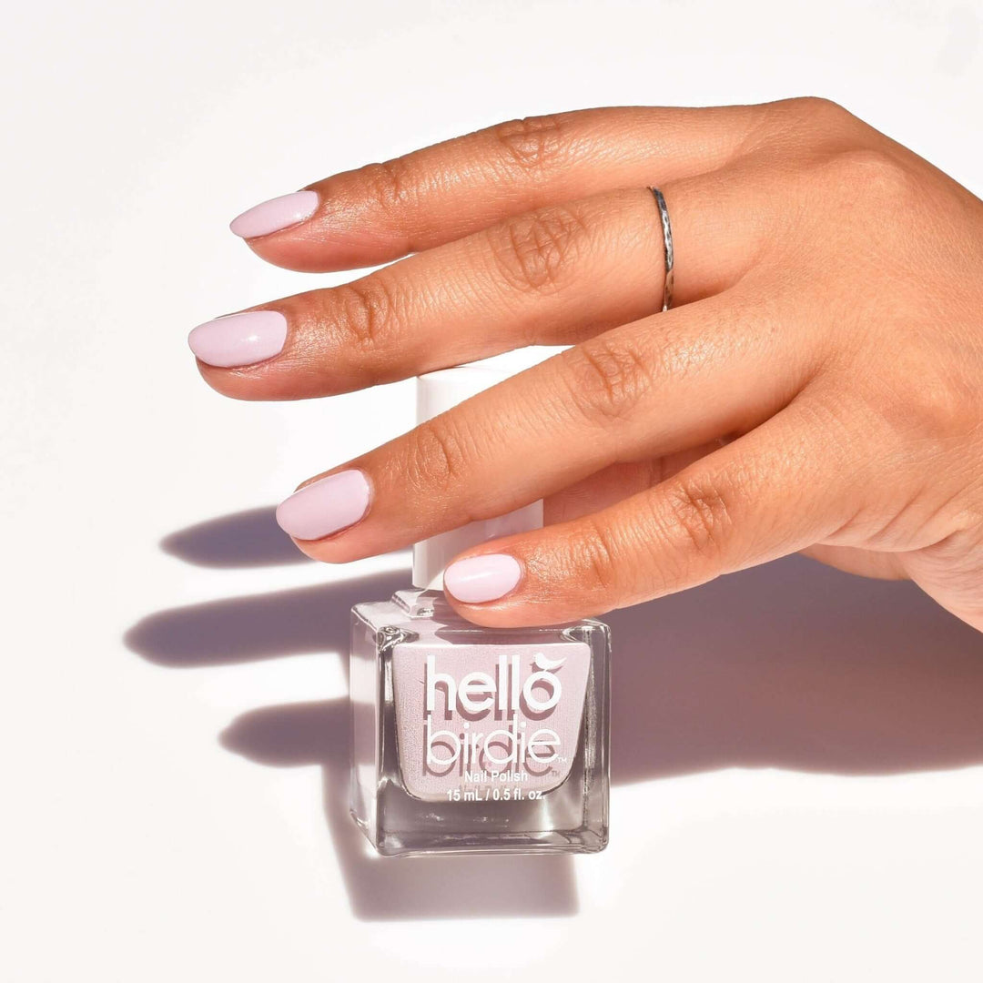 A close up of a mid toned hand comes into frame from the right side in front of a bottle of Hen Party nail polish from Hello Birdie in a light creamy pink. The nails have the same polish on and there is a silver ring on one finger. The bottle is shaped like a cube and against a matte white background with a hard light shadow beneath them.