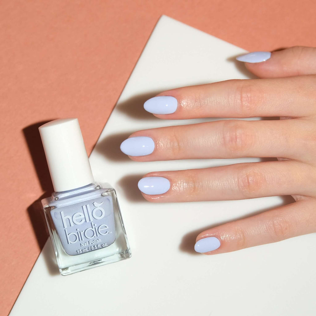 A close up of a light toned hand wearing Tweet Dreams nail polish from Hello Birdie which is a pale blue color. A bottle of the same polish is to the left of the hand and is cube shaped with a white cap and text. The hand is brightly lit in front of a cream and tan offset background.
