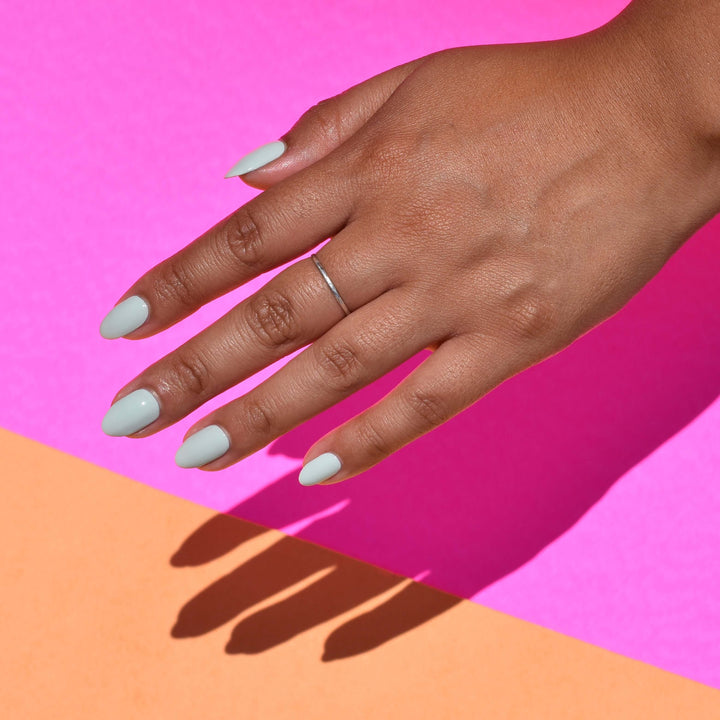 A close up of a hand against a bright pink and tan paper background. The hand is brightly lit and casting a sharp shadow. Wingin' It nail polish from Hello Birdie is painted on the nails in a light sage hue and the middle finger has a thin silver ring.