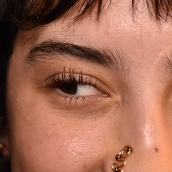 Close-up of a person with a nose ring and hoop earrings against a purple background and a side view of her lashese after a lash lift