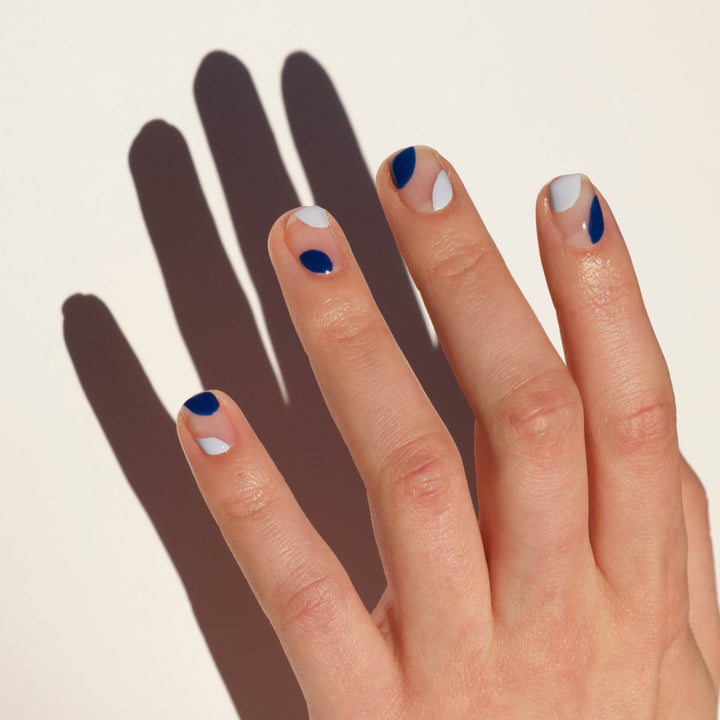 A Close up of a model's hand with light skin tone and negative space nail art on her nails in light blue and royal blue. A hard light shadow of the hand falls on a white background.