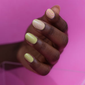 Close up of model's hands wearing Hello Birdie Nail Polish in a light lime green and light peachy pink combined on two nails of nail art  in a swirl line pattern. The hand is placed infront of a soft bubblegum pink background with only the nails in focus.