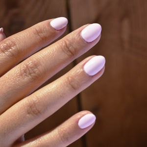 A close up of a model's fingers wearing Lovebirds nail Polish from Hello Birdie in a light pink hue with medium skin tone and a deep caramel colored wood background.