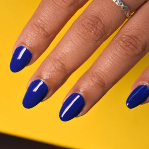 A close up of a model's fingers wearing Myna Your Own Business nail polish from Hello Birdie. The polish is royal blue and glossy and the background is a bright crayon yellow.