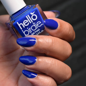 Close up of a model's hand holding a bottle of Myna Your Own Business nail polish from Hello Birdie in a royal blue hue. The polish is on the nails and the skin tone is medium. The background is out of focus and dark grey.