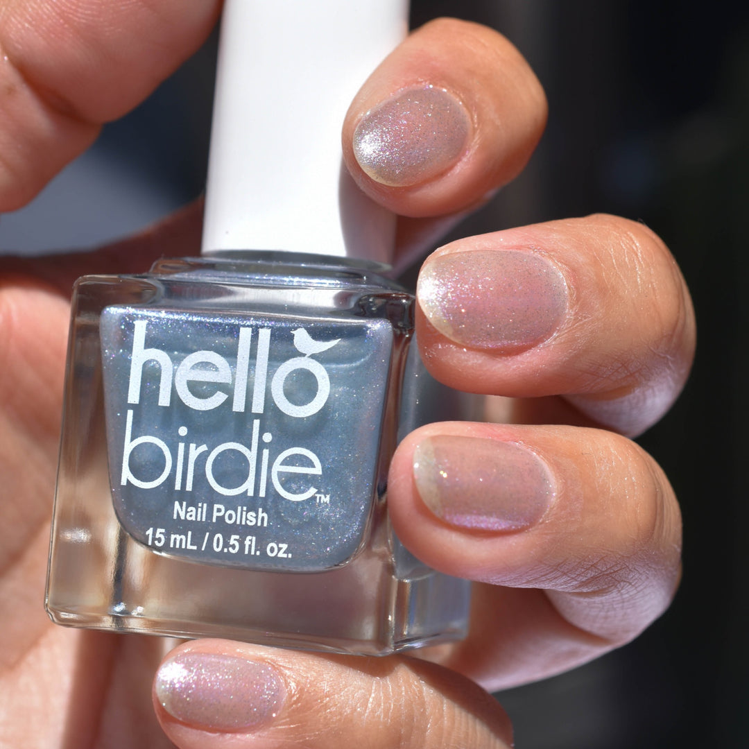 A close up of a bottle of Nightingale nail polish from Hello Birdie. The bottle has silvery blue translucent glitter in it and is painted on the nails of the hand. The hand is illuminated from the front and below.