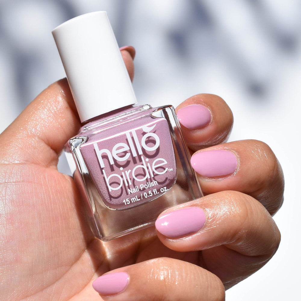 A close up of a mid toned hand holding a bottle of Owl You Need is Love nail polish from Hello Birdie in a light taupe purple hue. The bottle has white text and white cap and is cube in shape. The nails of the hand is painted with the same color and the background has soft shadows of leaves against matte white.