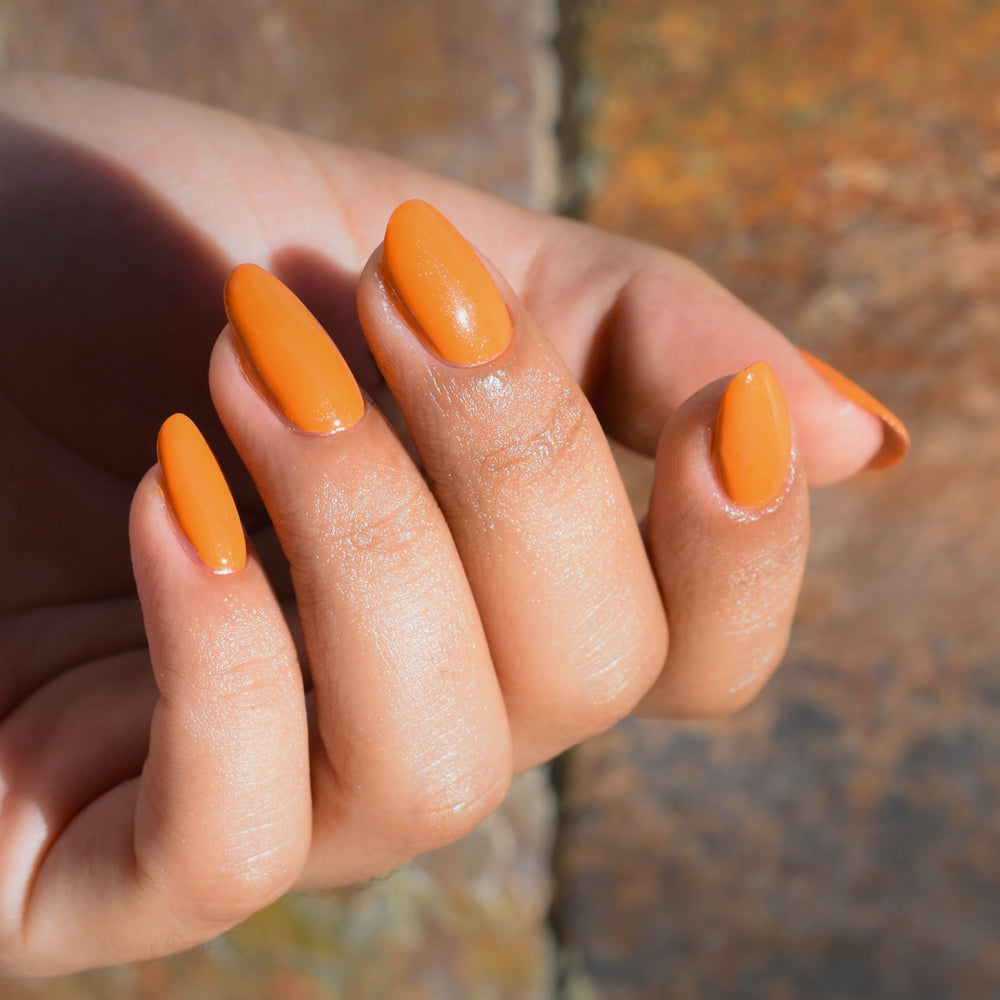 A close up of a model's fingers wearing Phoenix nail polish from Hello Birdie which is a deep orange hue. The background is out of focus warm colored stone tiles.