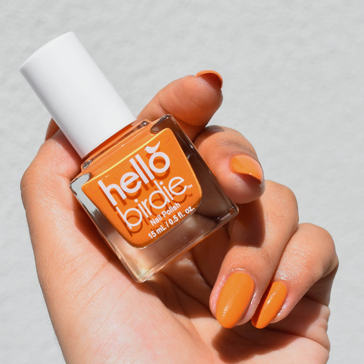A close up of a medium toned hand holding a bottle of Phoenix nail polish from Hello Birdie which is a deep orange hue. The polish is on the nails and the background is a soft white wall