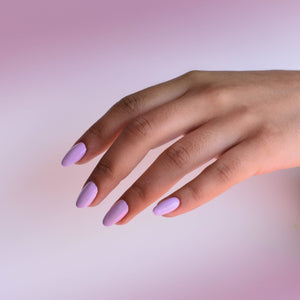A medium toned hand floating in from the right wears Sweetie Bird nail polish from Hello Birdie which is a pinky lavender hue. The background is a cloudy blush color.