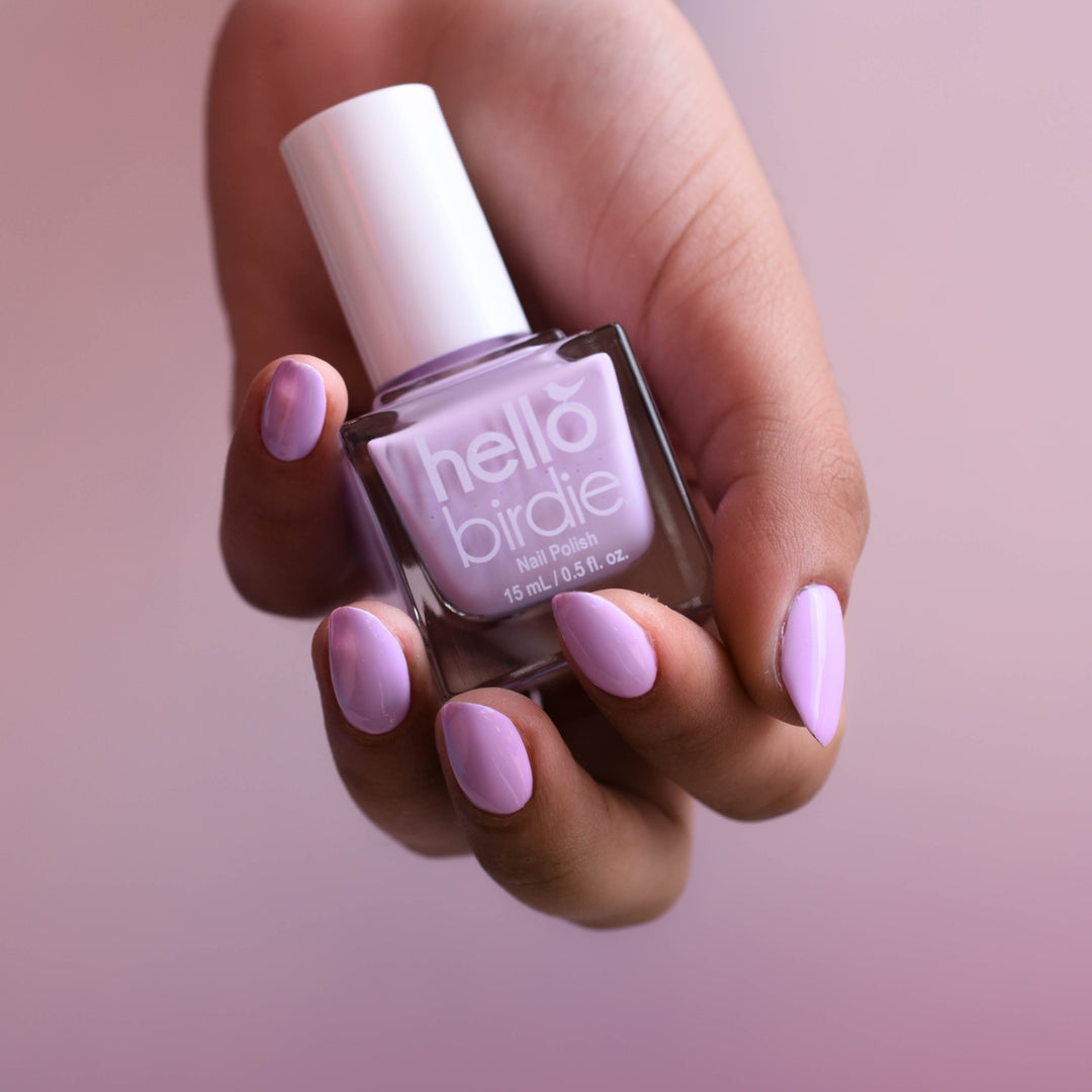 A close up of a medium toned hand clutching a bottle of Sweetie Bird nail polish from Hello Birdie which is a pinkish lavender hue. The polish is painted on the nails and the background is a soft cloudy blush color.