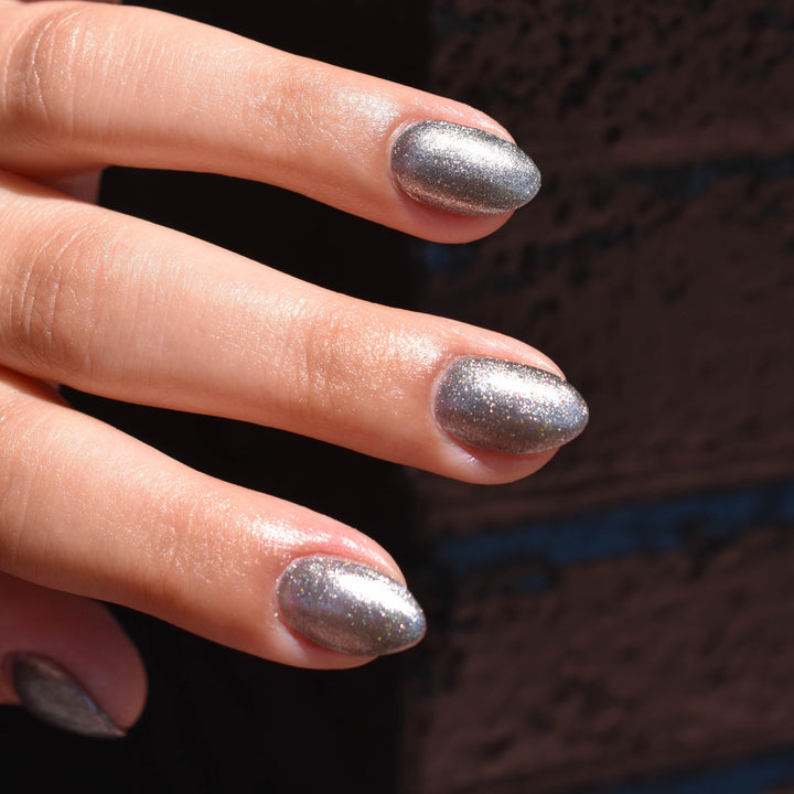 Four fingers are featured wearingSpace Bird nail Polish from Hello Birdie which is a charcoal metallic hue. The hand is brightly lit, the nails are shiny and sparkling while the background is a glossy black brick corner.