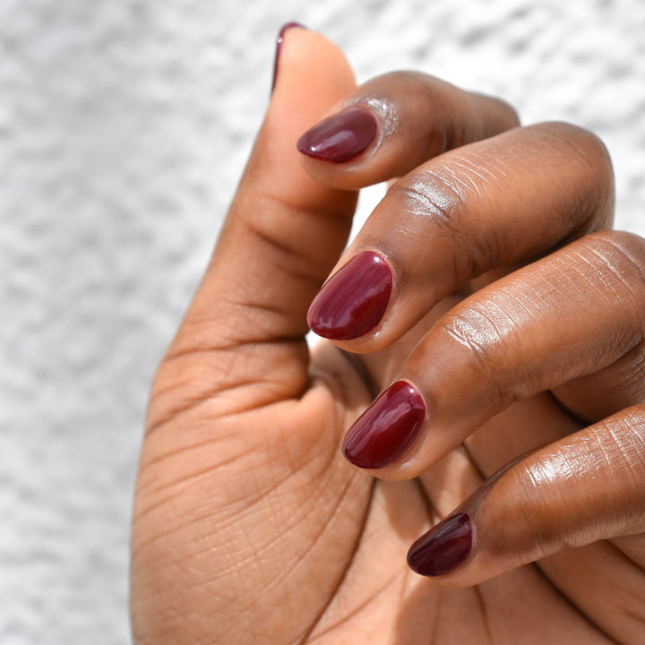 A model's hand showing Hello Birdie Nail Polish in Bird of Prey on her nails with a deep skin tone. Bird of Prey is  a deep Maroon color.