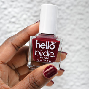 There is a model's hand with deep skin tone holding a bottle of Hello Birdie Nail Polish in Bird of Prey. The color is a deep maroon and is on the model's nails.