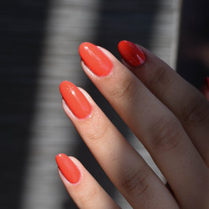 Close up of model's hand with medium skin tone wearing Cheep Thrill Nail Polish from Hello Birdie which is a poppy red. A beam of light highlights the fingertips and reveals the silver metallic background.