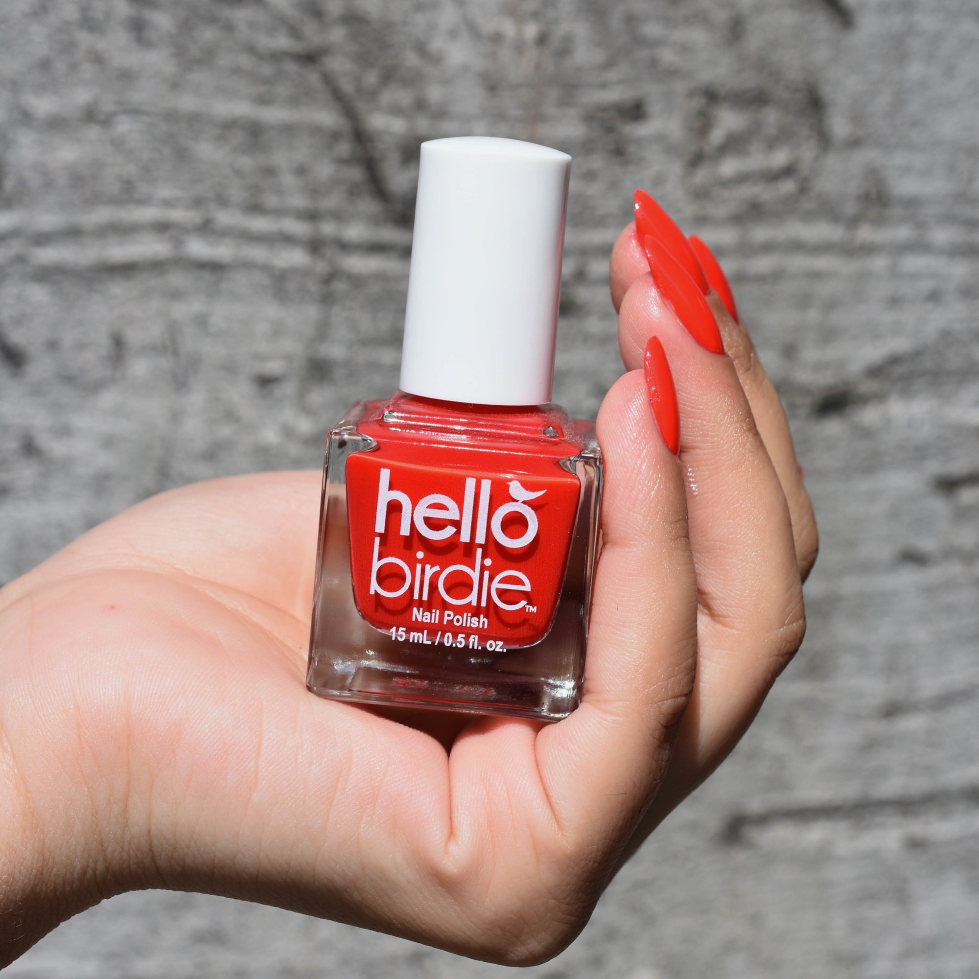 A model is holding a bottle of Cheep Thrill from Hello Birdie Nail Polish. The color is a poppy red and is in the bottle and painted on the model's hand which has a medium skin tone. The background is a grey bark.