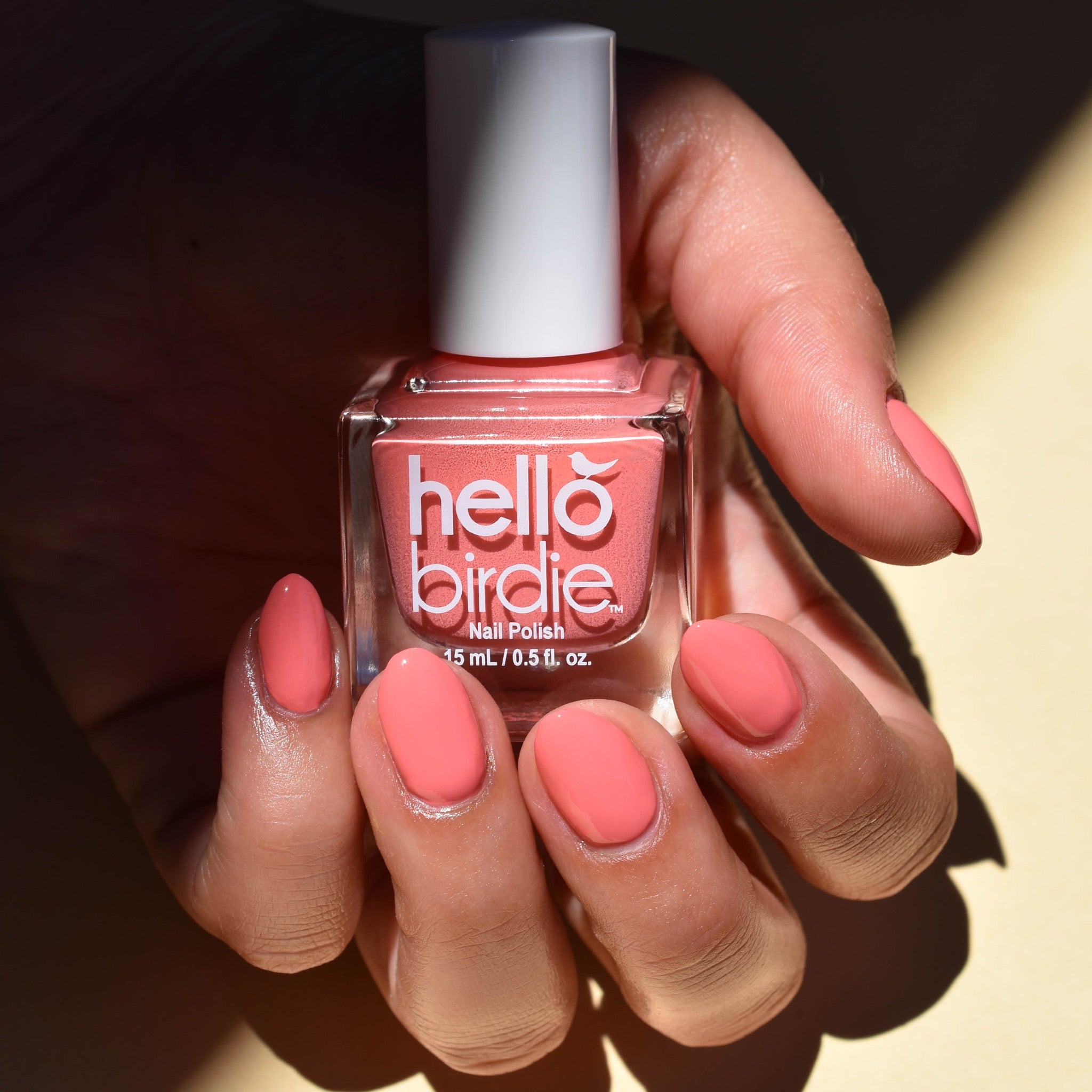 A close up of a mid toned hand is holding a bottle of Cupid Sparrow nail polish from Hello Birdie which is a deep blush hue. The bottle is cube shaped and has a white cap and text. The nails are painting with the same polish. A beam of light illuminates the bottle and hand and the background is a cream color.