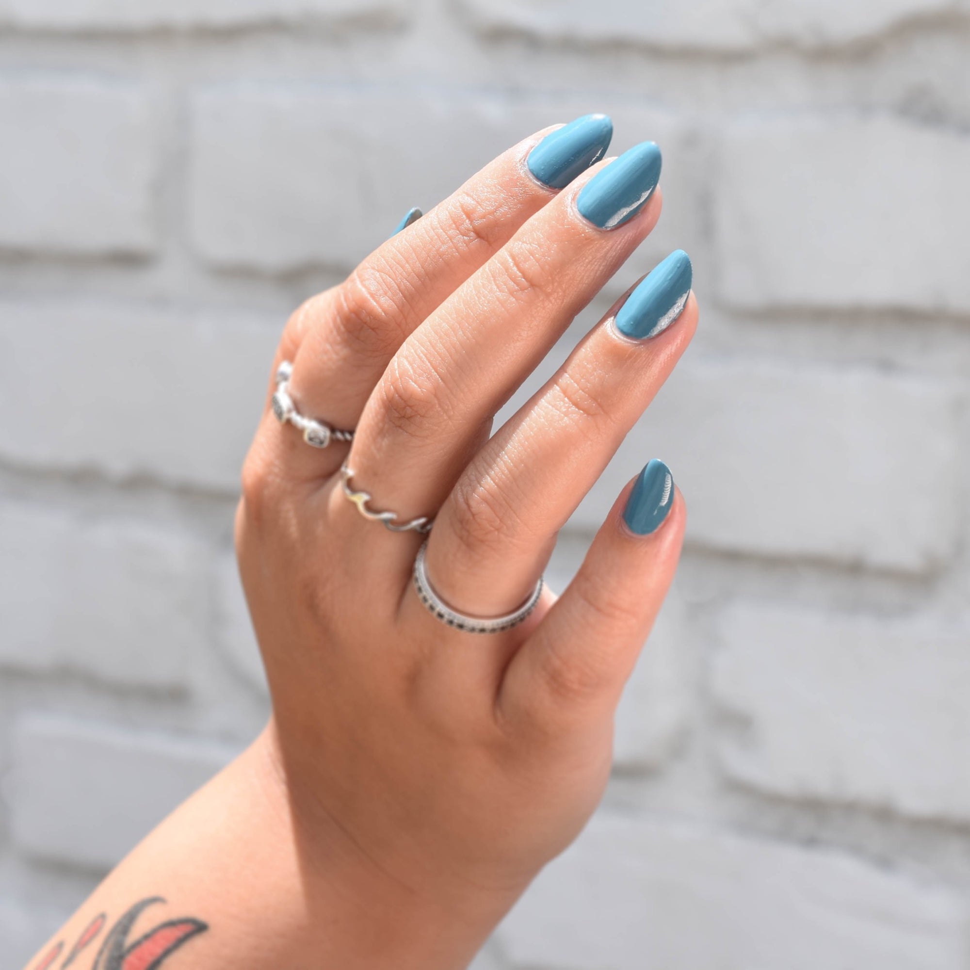 A Model's hand with medium skin tone in daylight wearing Eggsistential Nail Polish from Hello Birdie which is a muted blueish teal. Model is wearing three silver rings and a glimpse of her tattoo on her arm is shown. The background is a painted white brick.