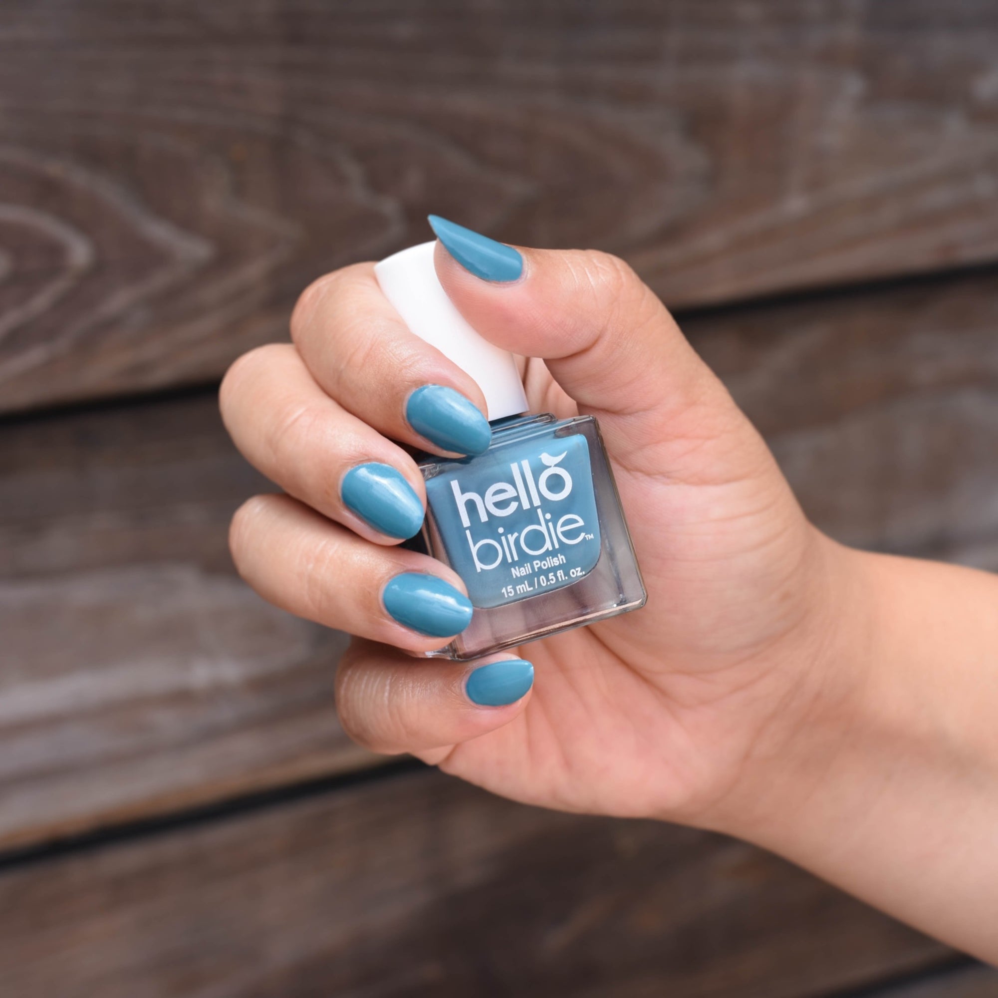 A model's hand is shown holding a bottle of Hello Birdie's Eggsistential nail polish. She has the color painted on her nails and it is a muted blueish teal.  The background shows horizontal wood panels.