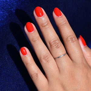 A close up of a mid toned hand coming within frame from the lower right corner wearing Emuse Me nail polish from Hello Birdie. The polish is a bright classic red hue and the model has a silver ring. The Background is a dark royal blue velvet and shimmery.