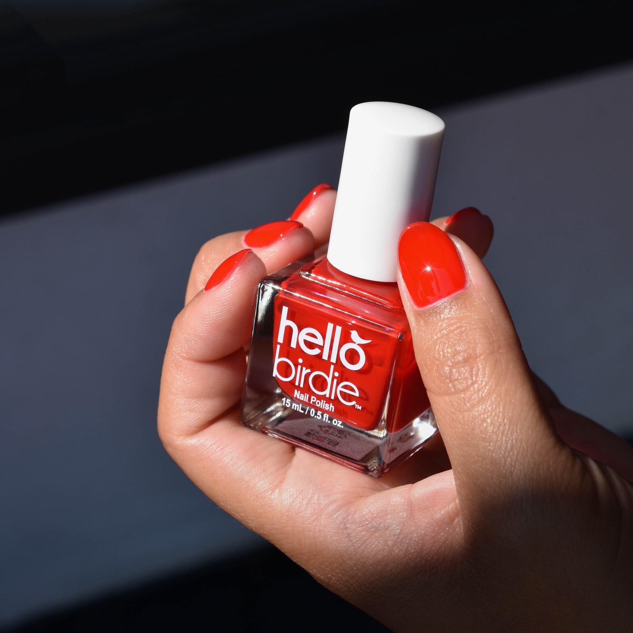 A close up of a hand comes into view from the lower right corner holding a bottle of Emuse Me nail polish from Hello Birdie which is a bright classic red hue. The model is also wearing the polish and the nails are glossy. The bottle has a cube shape, white cap, and white text with logo. A beam of light illuminates the hand within a black space.
