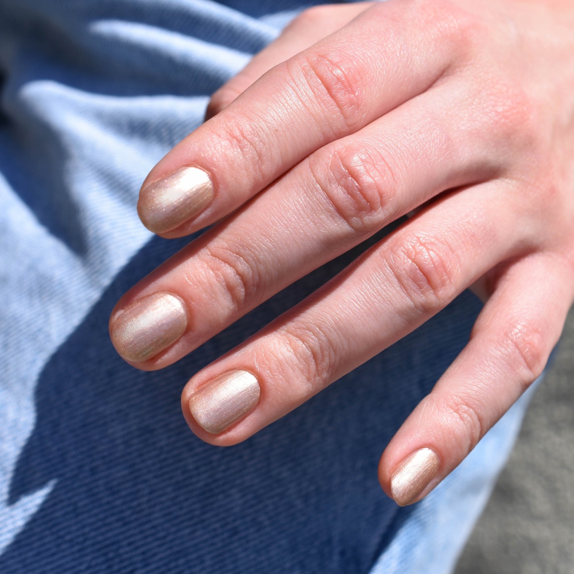 A close up of a light toned hand wearing Feathered nail polish from Hello Birdie which is a pale bronze color. The hand comes from the upper right hand corner and the background are light denim jeans.