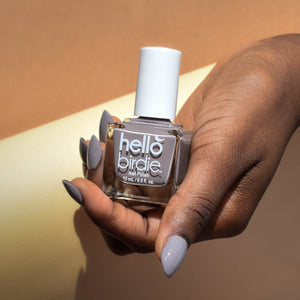 A model is holding a bottle of For Flock's Sake nail polish from Hello Birdie. The model is wearing the polish in a muted taupe gray on her nails and has a deep skin tone.  The background is cut diagonally with the top half in a caramel brown and the bottom half in a sandy yellow.