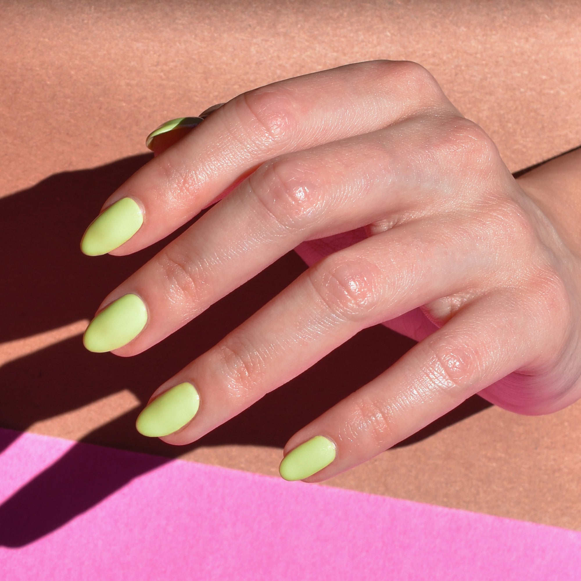 A light toned hand is featured up close with Good Migrations nail polish from Hello Birdie painted on the nails in a light lime green hue. The hand is brightly lit against a tan and pink paper background.