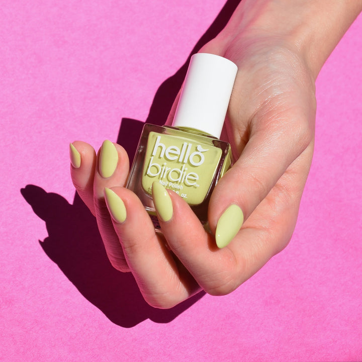 A close up of a light toned hand comes into frame from the upper right hand corner holding a bottle of Good Migrations nail polish from Hello Birdie in a light lime green color. The bottle has a cube shape and white cap and text and the same polish is on the nails. The hand is brightly lit against a pink paper backdrop