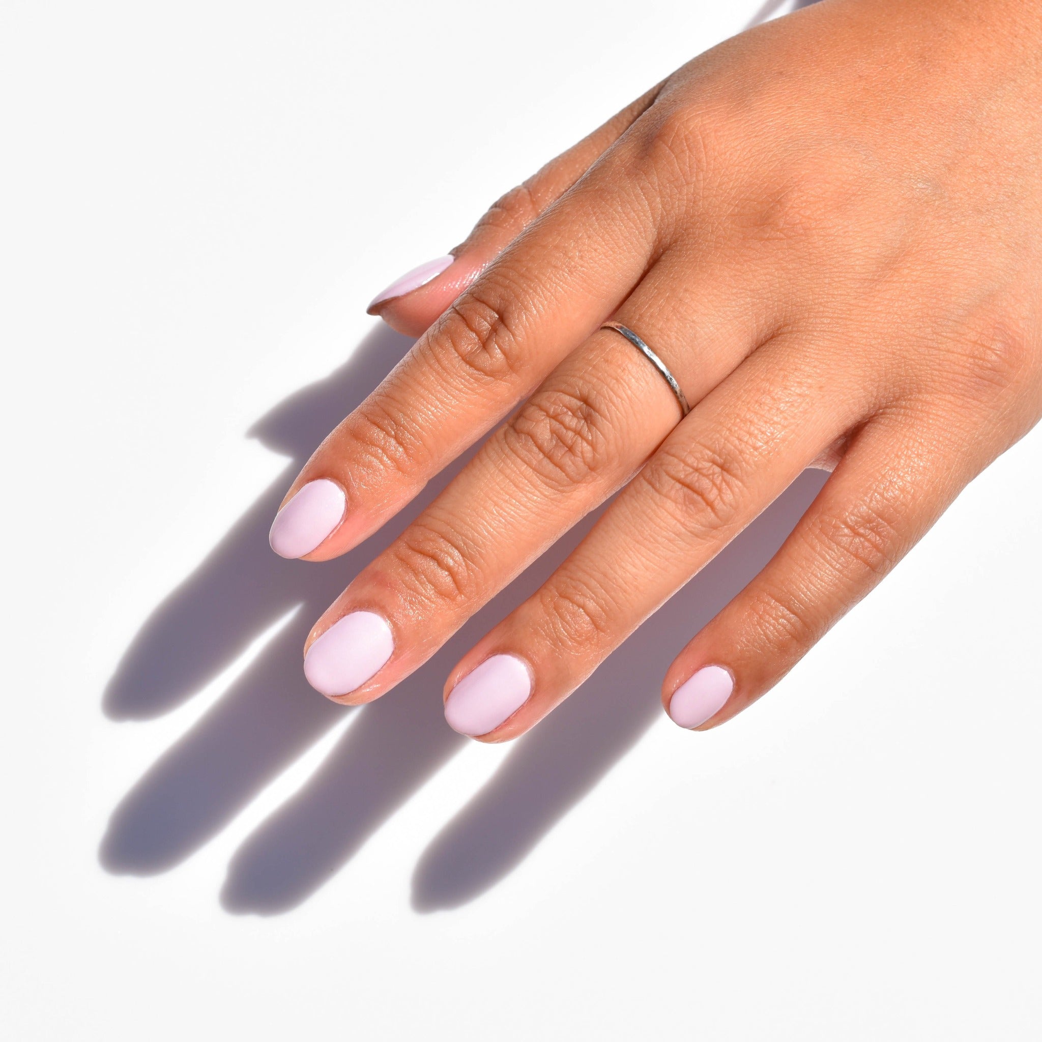 A close up of a mid toned hand comes in from the upper right corner wearing a silver ring and Hen Party nail polish from Hello Birdie in a creamy pink hue. The hand casts a hard light shadow diagonally against a matte white background.