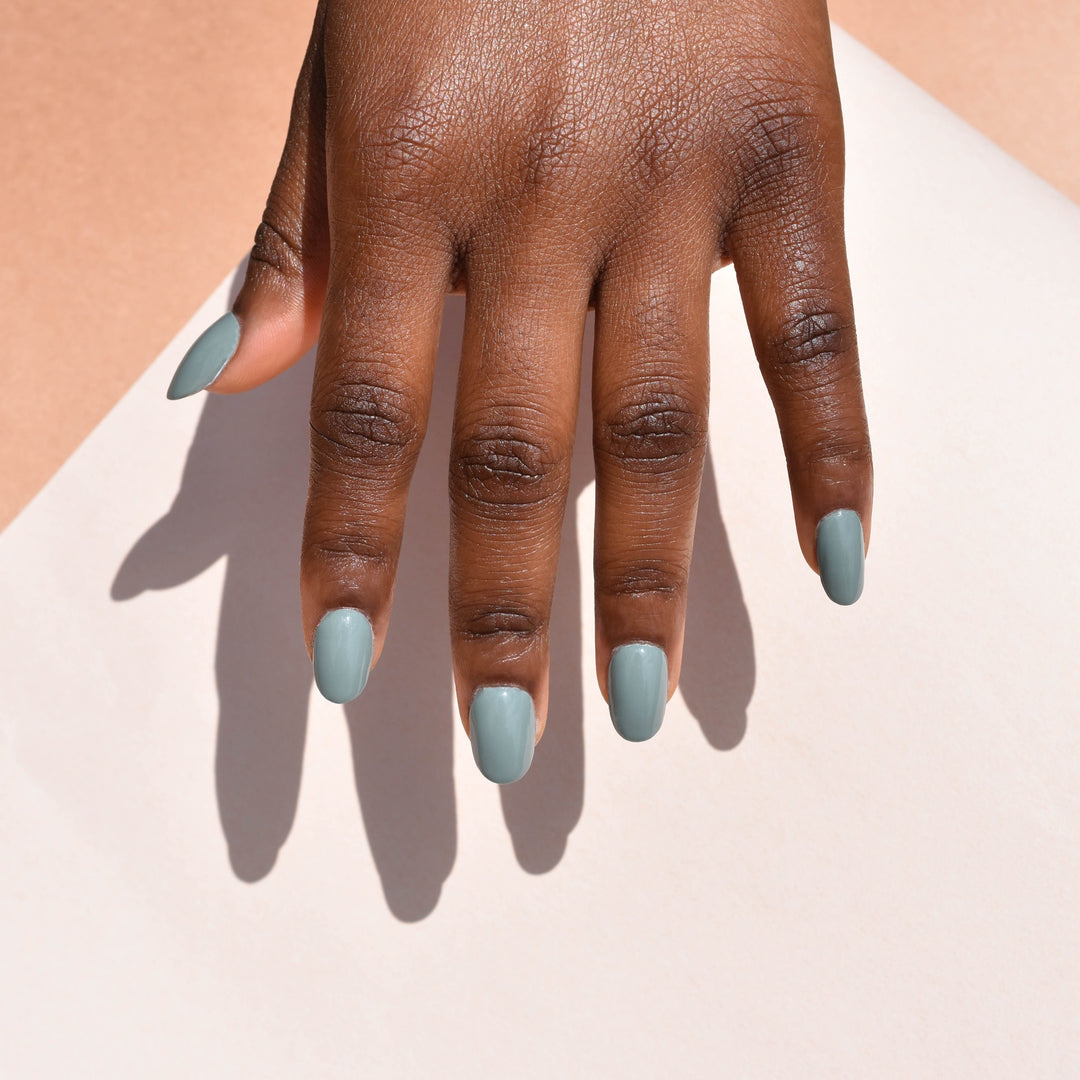 A model's hand coming from the top of the Frame wearing Impeccable nail polish from Hello Birdie in a medium sage green with a deep skin tone. The hand's  hard light shadow falls onto a background of eggshell and tan hues.