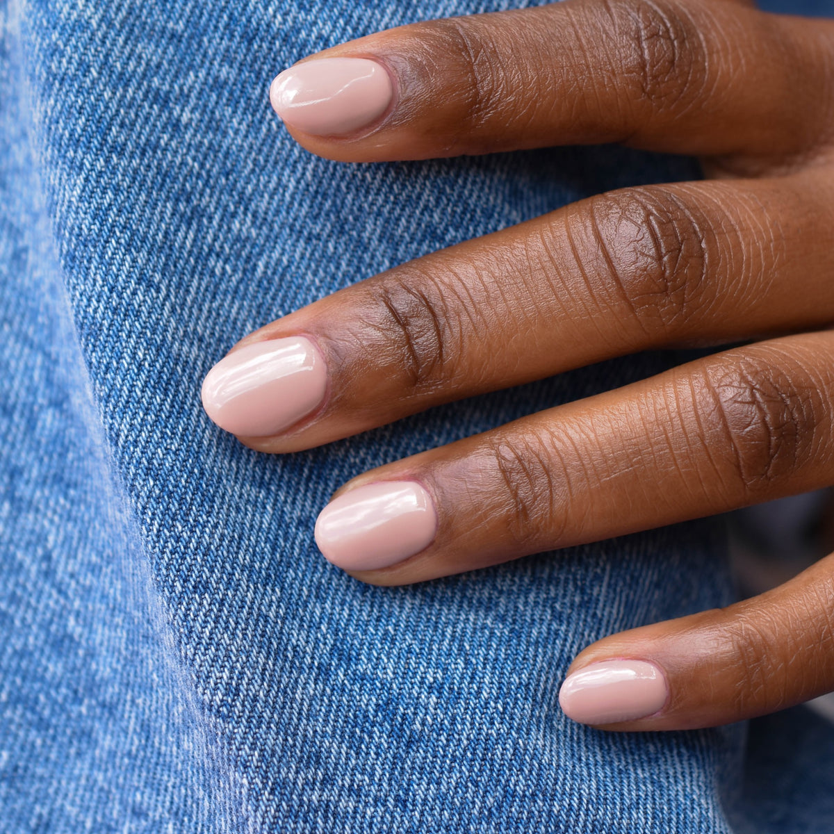 A close up of a model's hand showing fingers touching denim. It's goin' Down nail polish is painted on her nails  which is a light tan taupe hue and the skin tone is deep.