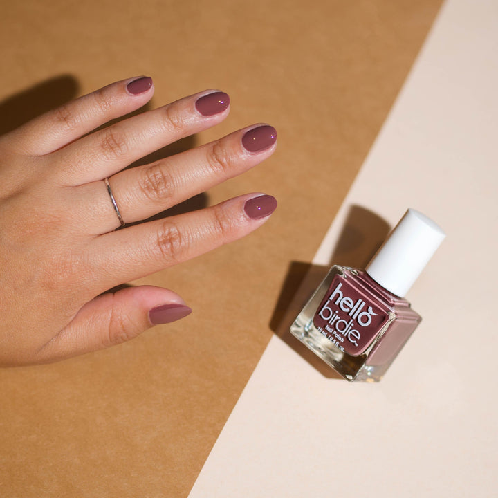 A mid toned hand comes within frame from the left wearing Wanna Be Nest to You nail polish from Hello Birdie with one finger having a silver ring. A bottle of the same polish floats to the right with a hard light shadow. The background is split with tan on the left side and cream on the right.