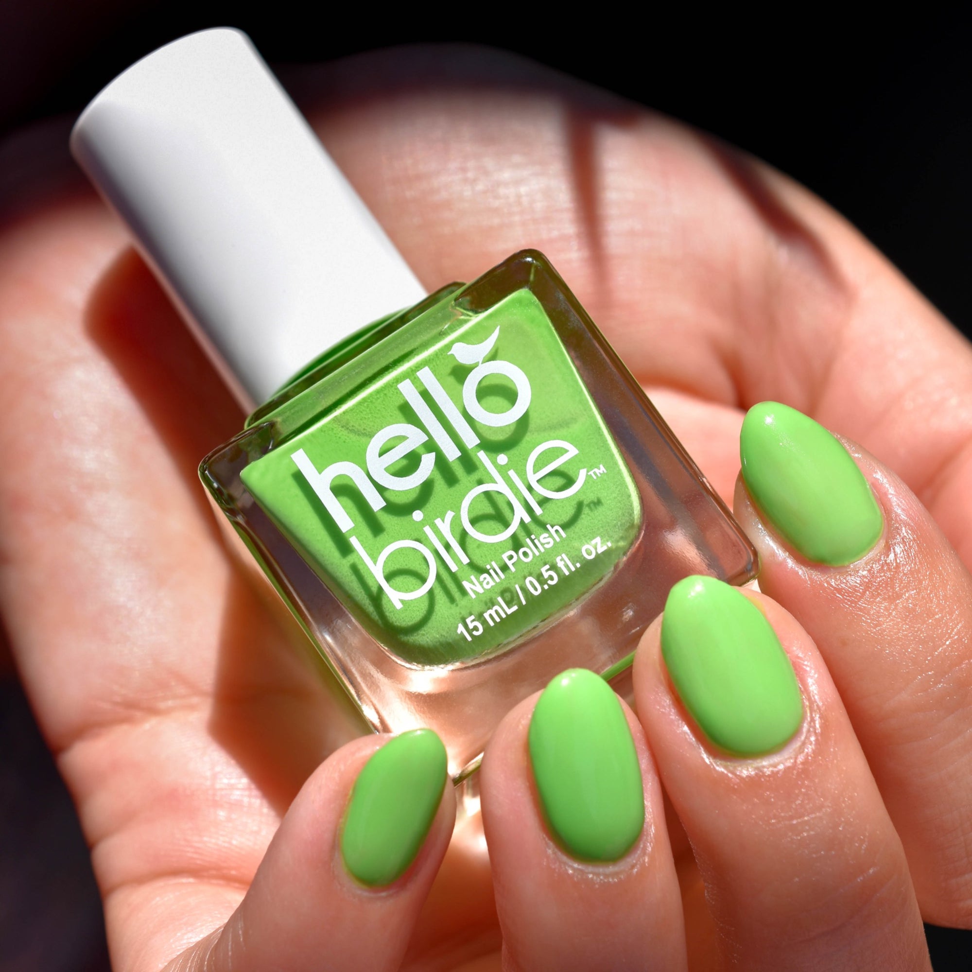 A close up of a mid toned hand clutching a bottle of Kiwi Nail Polish from Hello Birdie which is a lime green.  The nails of the hand are painted with the same color and the polish bottle has a cube shape and a white cap with white text. Dramatic sunlight highlights the hand with a leaf shadow over the palm and dark background.