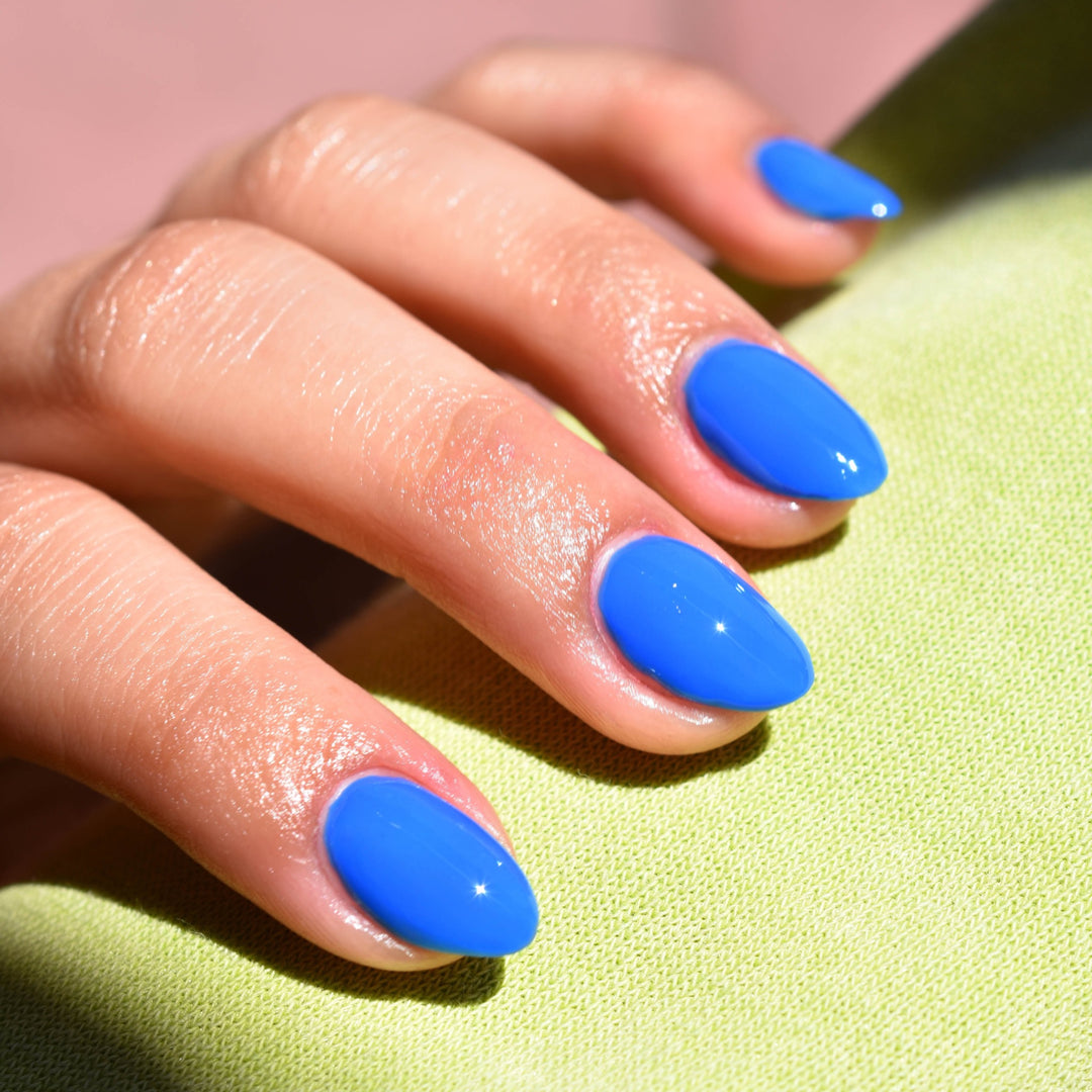 A close up of four fingers of a mid toned hand over light lime green cloth. A beam of light illuminates the fingers and the nails are painted with Mile High nail polish from Hello Birdie in a technicolor blue hue.