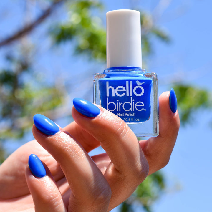 A close up of a hand holding up a bottle of Mile High nail polish from Hello Birdie, which is a technicolor blue hue. The bottle is shaped like a cube and the cap and text are white. The model's finger nails are painted with the same polish and in the background tree branches with green leaves are out of focus in the sun.