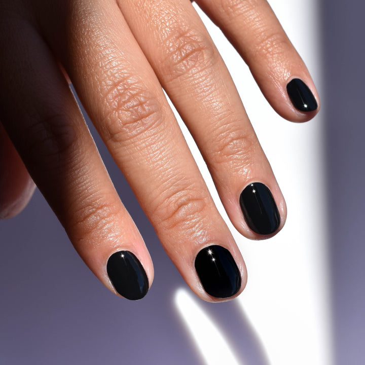 Close up of a model's hand with medium skin tone wearing Nevermore nail polish from Hello Birdie which is a deep black hue. A beam of light highlights the nails and reveals the white background and hard light shadows.