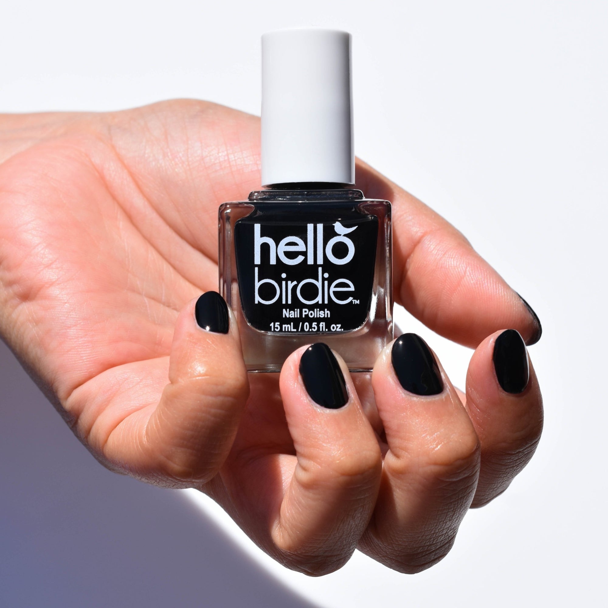 A Close up of a model's hand holding a bottle of Nevermore Nail Polish from Hello Birdie in the palm. The hand has a medium skin tone and the nails are painted with Nevermore, a deep black polish. Hard light falls on the hand with a white background and shadow underneath.