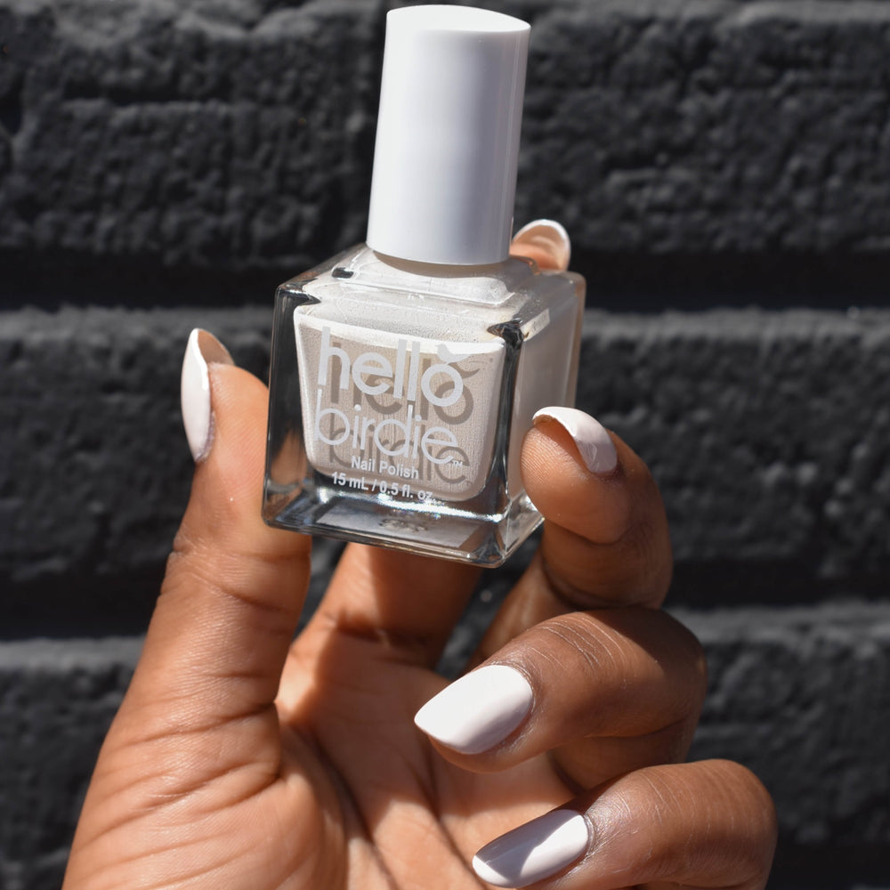 A Close up of a model's hand holding a bottle of No Egrets nail polish from Hello Birdie. The polish is an off-white nude and is painted on the nails of the hand that has a deep skin tone. Out of focus is a black brick background.