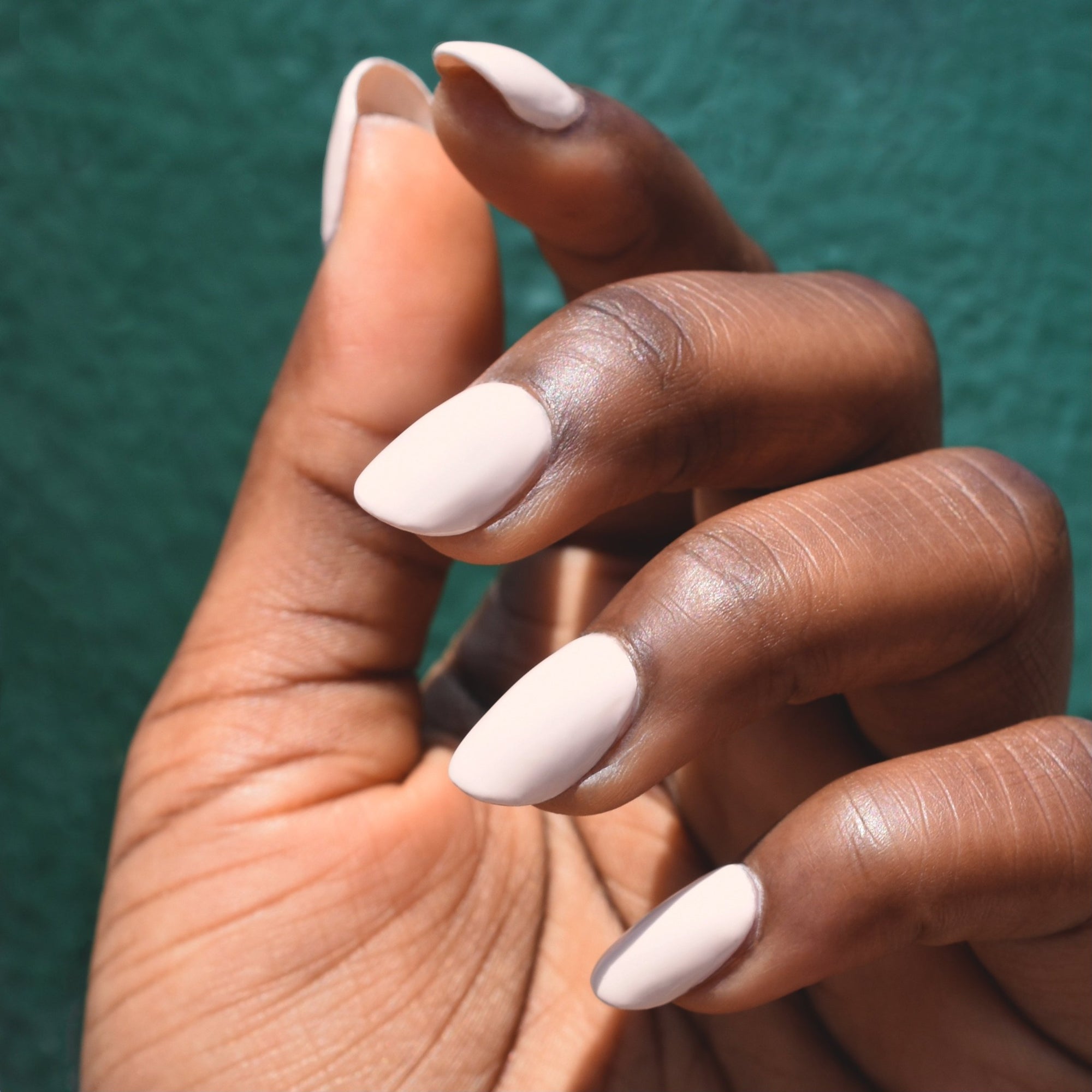 A model's hand is in front of a forest green wall. Her nails are painted with No Egrets nail polish from Hello Birdie which is an off-white nude and the skin tone is deep and lit with bright daylight.