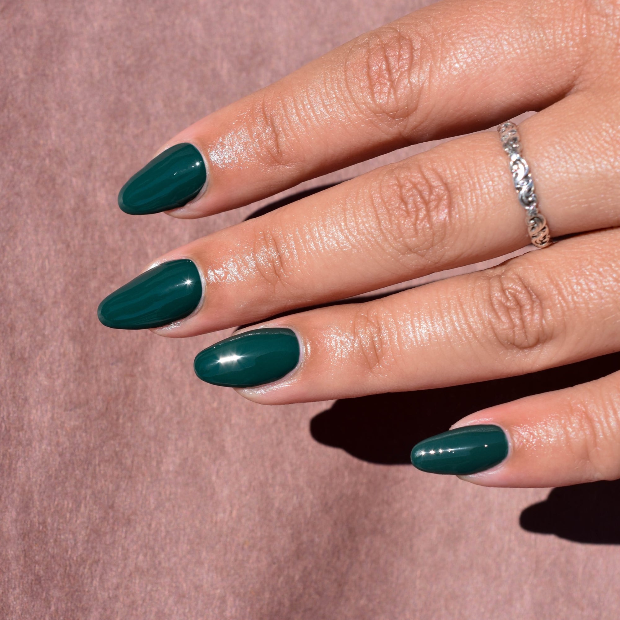 A close up of the fingers of a model with medium skin tone wearing No Harm No Fowl nail polish from Hello Birdie. The nails are a glossy dark green and one finger has a silver ring. Daylight sun creates a hard shadow of the hand on a papery light dusty rose background.