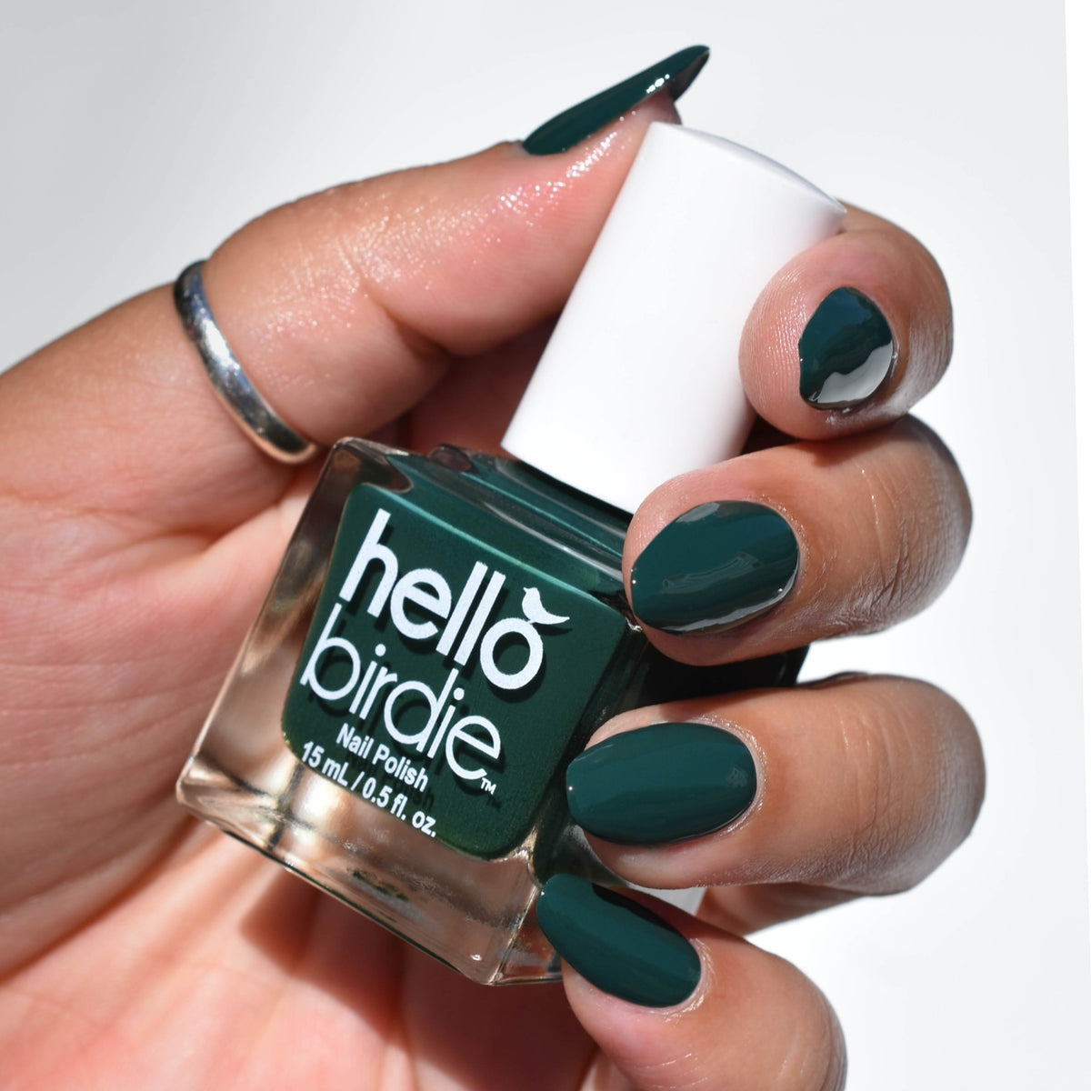 a Close up of a bottle of No Harm No Fowl nail polish from Hello Birdie. The polish is a dark green hue and is on the nails, the skin tone is medium. The thumb has a silver ring and the background is white
