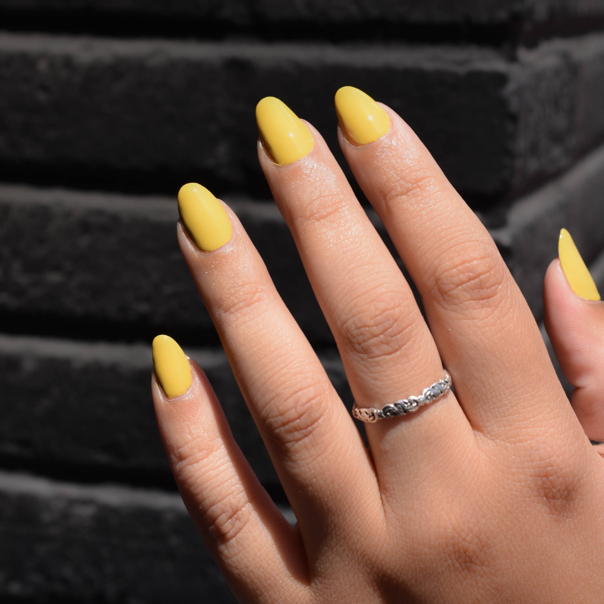 a close of of a model's hand wearing Over Easy nail polish from Hello birdie in a deep yellow hue. One finger has a silver ring and daylight illuminates it against an out of focus black brick corner background.