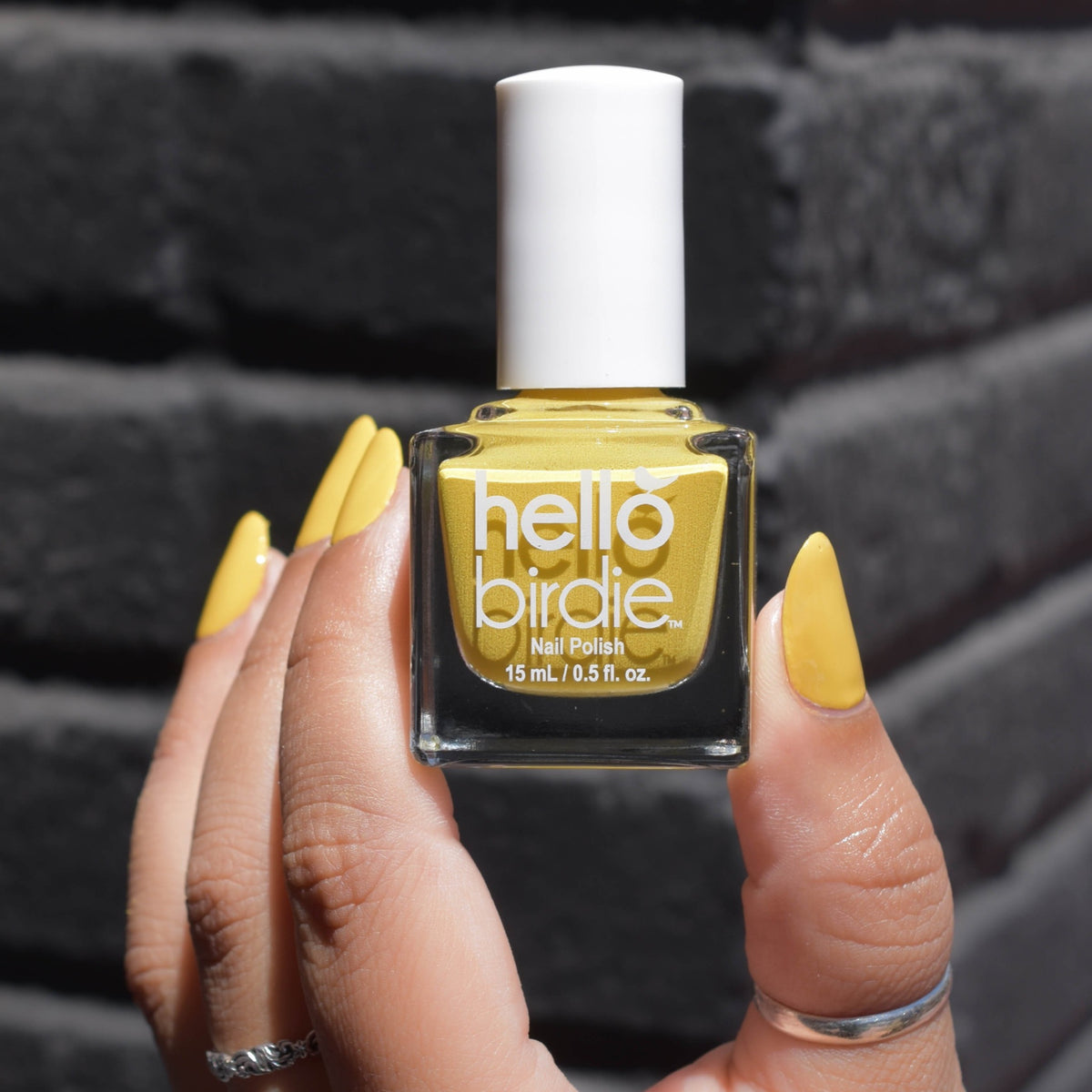 A close up of a model's medium toned fingers holding a bottle of Over Easy nail polish from Hello Birdie which is a deep yellow hue. The polish is painted on the nails and two fingers have silver rings. The background is a black brick corner which is out of focus.