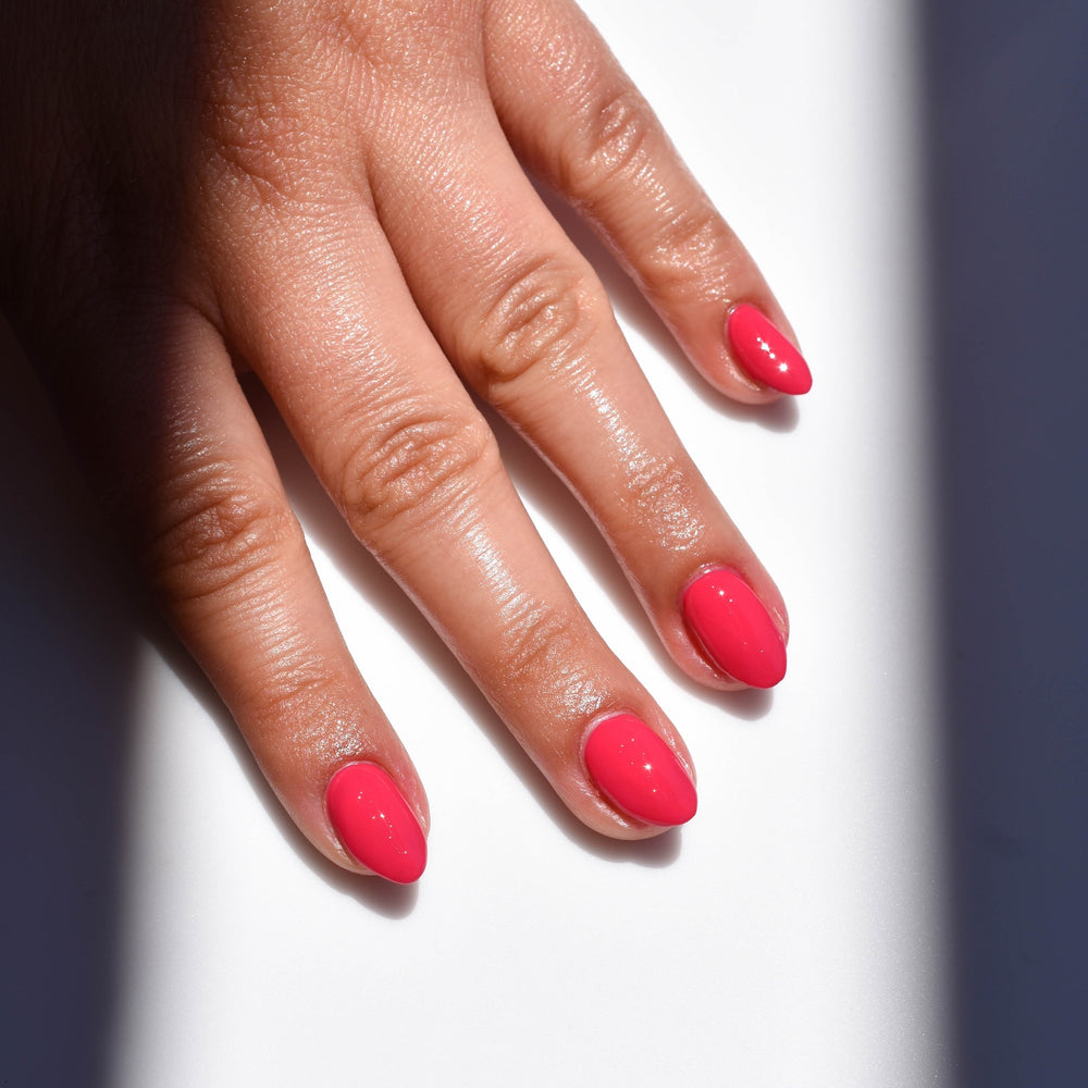 A beam of light falls on a mid toned hand highlighting the Pink Flamingos nail polish from Hello Birdie on the nails. The polish is a hot pink and the background is white with the right and left sides in shadow.