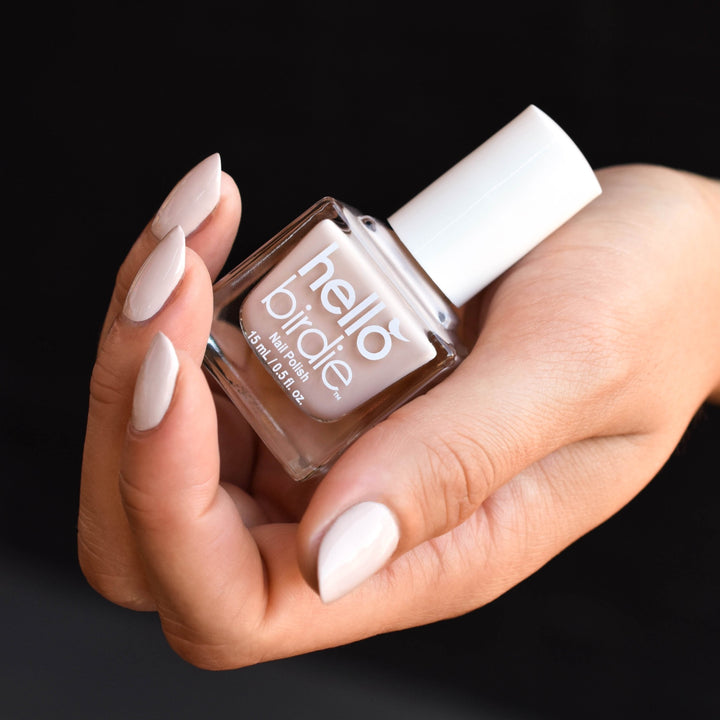 A close up of a hand cradling a bottle of Powder Down nail polish from Hello Birdie which is a neutral beige hue. The nails are painted with the same color and the bottle is cube shaped with white cap and text. The background is in shadow.