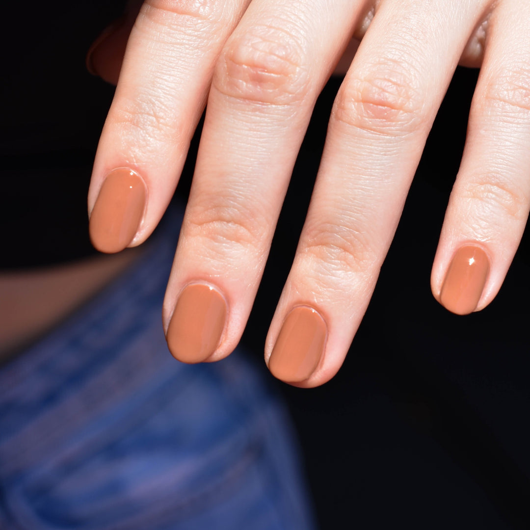 A hand comes in from the upper right hand corner, brightly illuminated. The nails are painted with Quail nail polish from Hello Birdie which is a medium caramel color. The background shows the models midsection, black shirt and jeans in shadow.