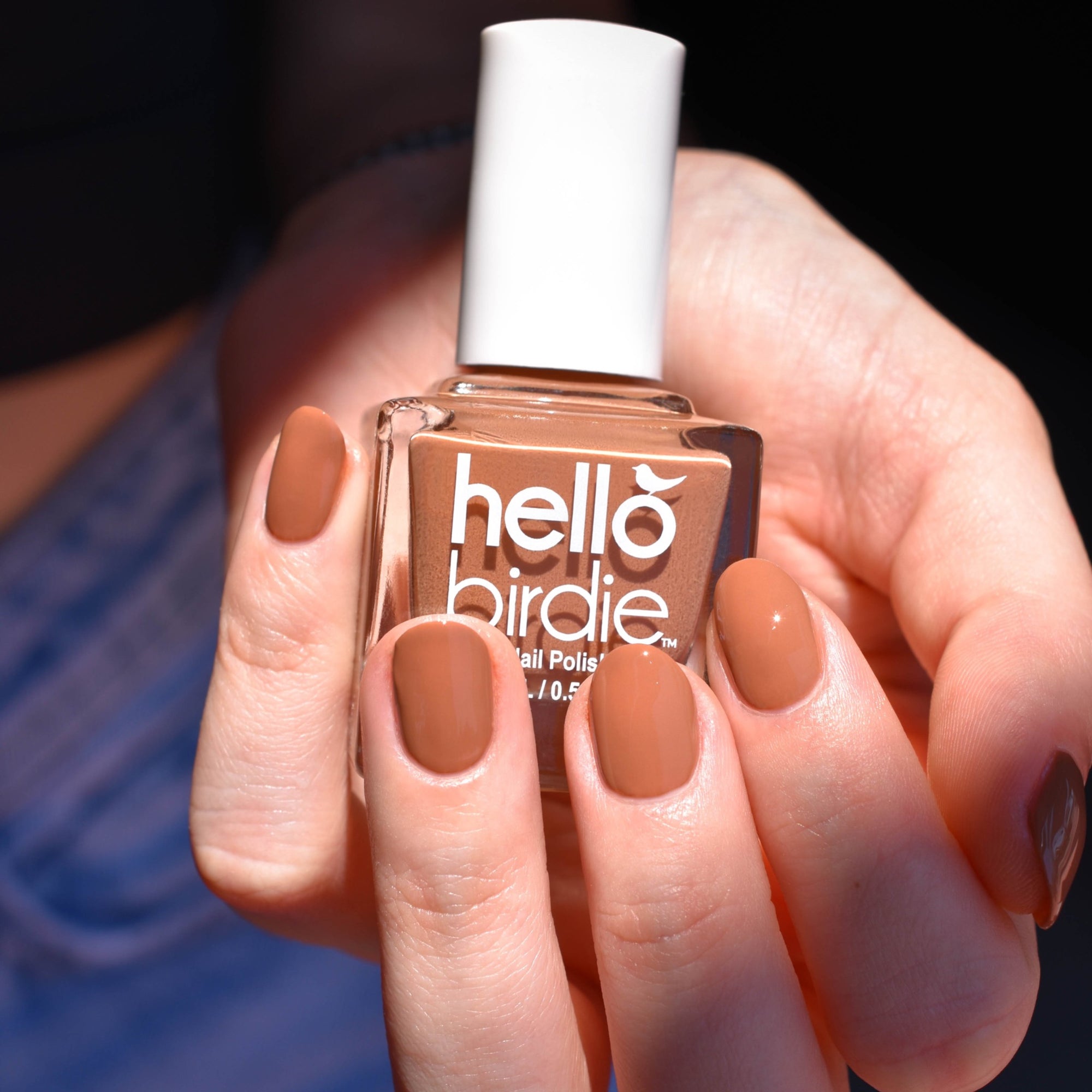 A close up of a light toned hand is illuminated by a beam of light. The hand is cradling a bottle of Quail nail polish from Hello Birdie which is a medium caramel color. The bottle is cube shaped with a white cap and white text. The nails are painted with the same polish and the background is the model's midsection with black shirt and jeans in shadow.