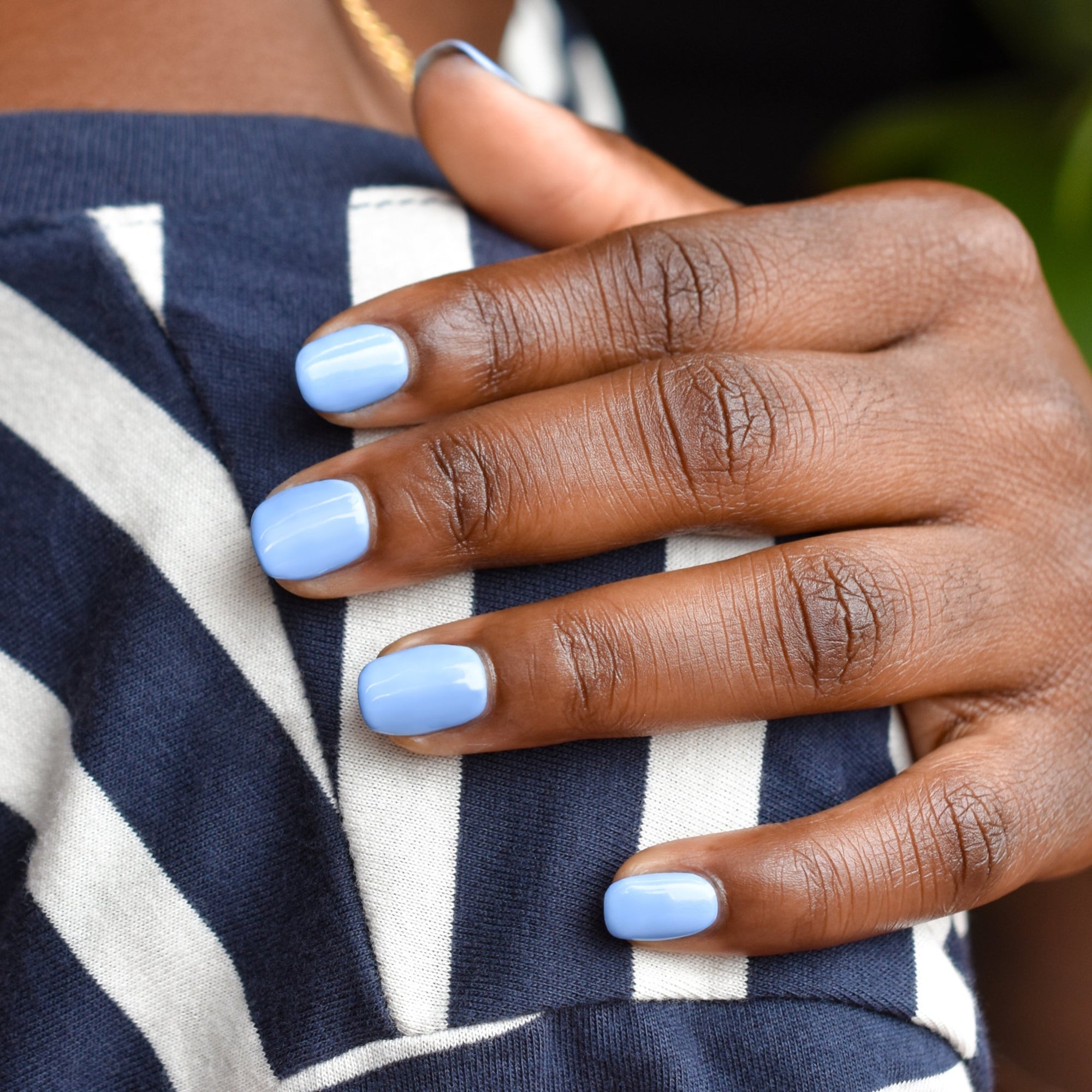 A close up of a hand on a shoulder wearing So Fly nail polish from Hello Birdie which is a sky blue color. The hand rests on a navy and white striped shirt and greenery is out of focus in the background.